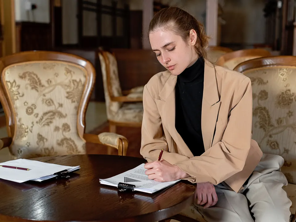 Vrouw in beige blazer tekent documenten aan een ronde houten tafel tussen klassieke fauteuils